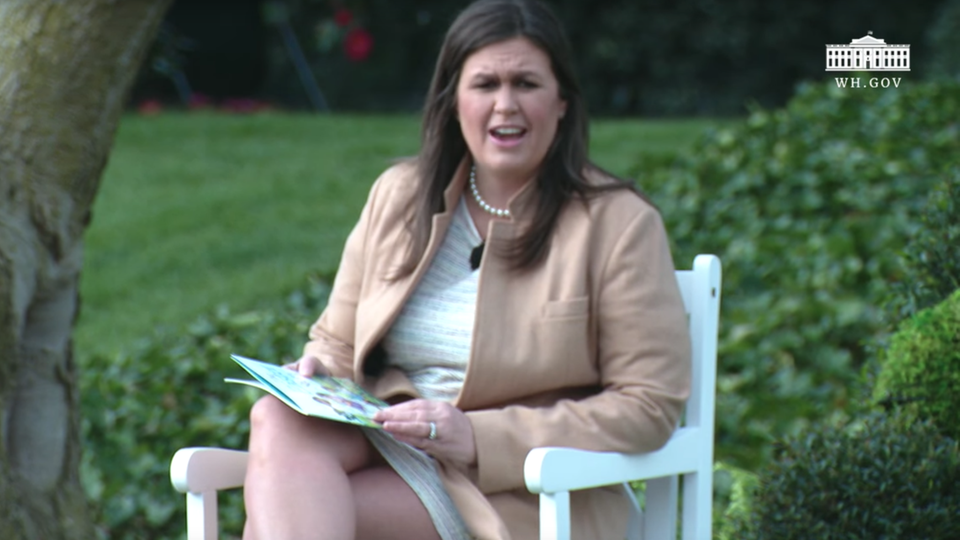 Sarah Huckabee Sanders sitzt auf einem weißen Gartenstuhl im Garten des Weißen Hauses. Auf dem Schoß hat sie ein Kinderbuch.