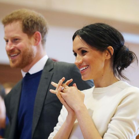 Megah Markle steht im weißen Wollpulli neben Prinz Harry und lächelt