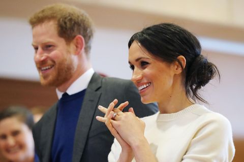 Megah Markle steht im weißen Wollpulli neben Prinz Harry und lächelt