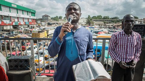 Ein Mann steht mit Mikrofon in der rechten und Bibel in der linken Hand auf dem größten Marktplatz in Accra.