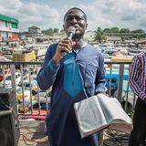 Ein Mann steht mit Mikrofon in der rechten und Bibel in der linken Hand auf dem größten Marktplatz in Accra.