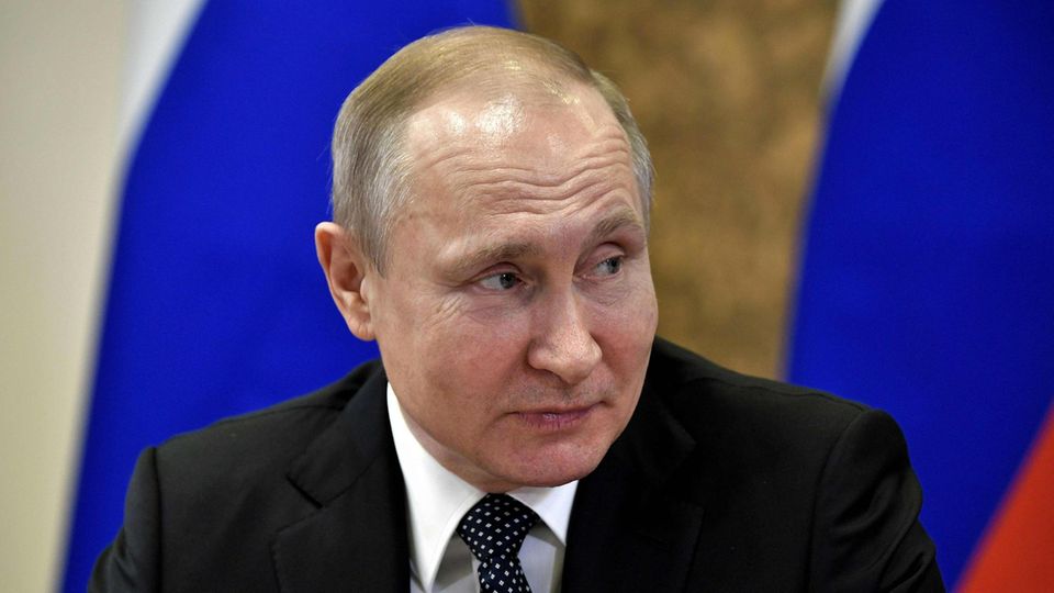 Mit einer russischen Flagge im Hintergrund sitzt Wladimir Putin an einem Tisch und schaut lächelnd zur Seite