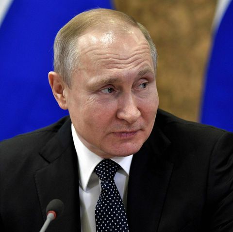 Mit einer russischen Flagge im Hintergrund sitzt Wladimir Putin an einem Tisch und schaut lächelnd zur Seite