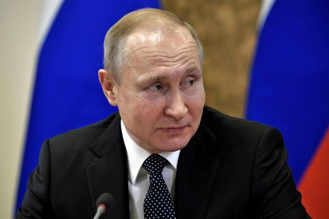 Mit einer russischen Flagge im Hintergrund sitzt Wladimir Putin an einem Tisch und schaut lächelnd zur Seite