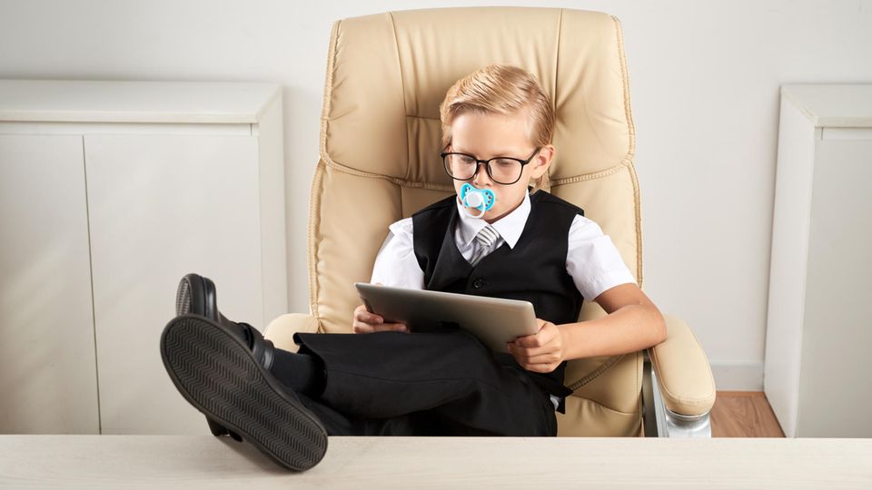 Ein Junge sitzt in einem Chefsessel und liest – mit Schnuller im Mund