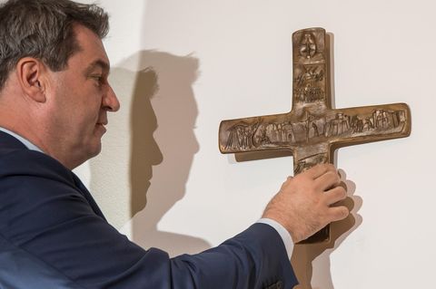 Bayerns Ministerpräsident Markus Söder steht vor einer Wand und hängt ein Kreuz auf. Er ist im Profil zu sehen und trägt Anzug
