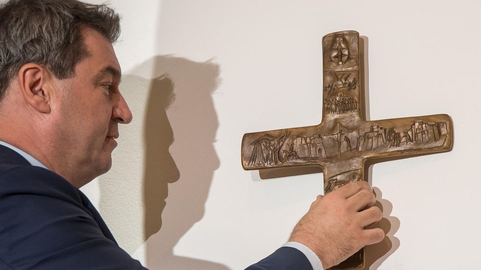 Bayerns Ministerpräsident Markus Söder steht vor einer Wand und hängt ein Kreuz auf. Er ist im Profil zu sehen und trägt Anzug