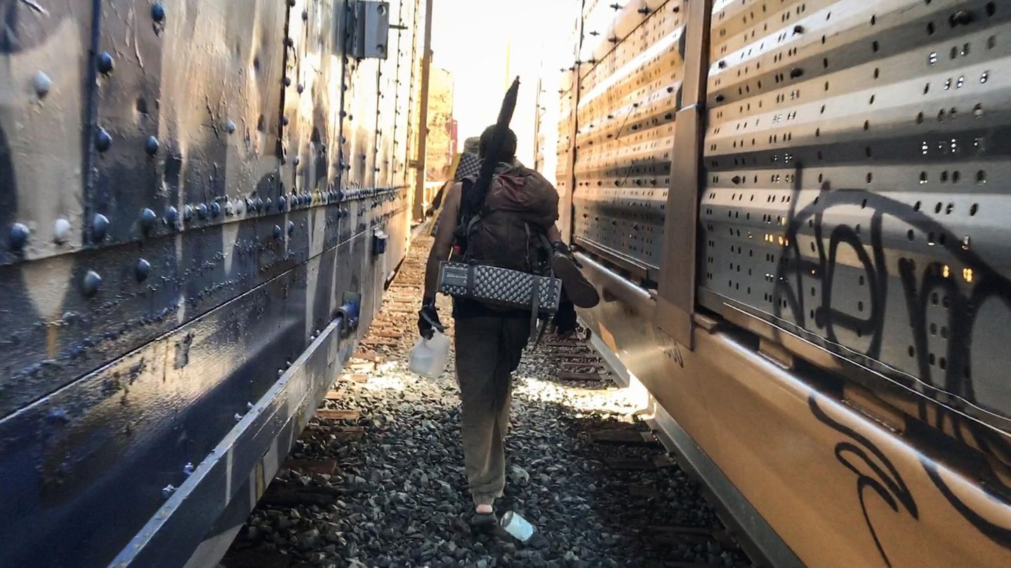 Ben läuft vor der Fotografin zwischen zwei stehenden Zügen, den Regenschirm am Rucksack. Wenn sie losfahren, kann es gefährlich werden