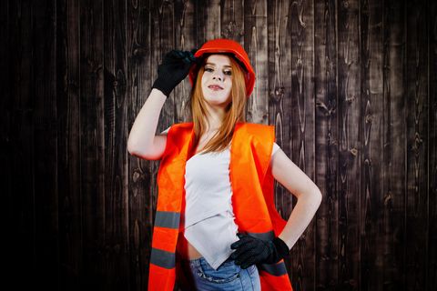 Rothaarige Frau in bauchfreiem Top, Hotpants, orangener Warnweste und Schutzhelm