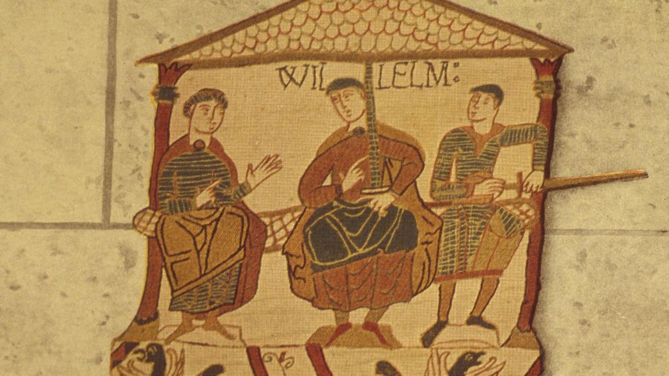 Ein Bild auf dem Teppich von Bayeux zeigt drei sitzende Männer in einem Haus. Über dem mittlerem steht "Willelm" für Wilhelm