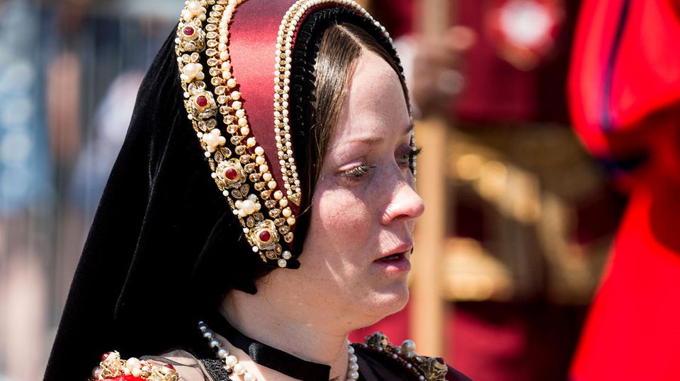 Anne Boleyn Eine dunkelhaarige Schauspielerin steht in einem dunkelroten, historischen Gewand mit einer passenden Haube in der Sonne