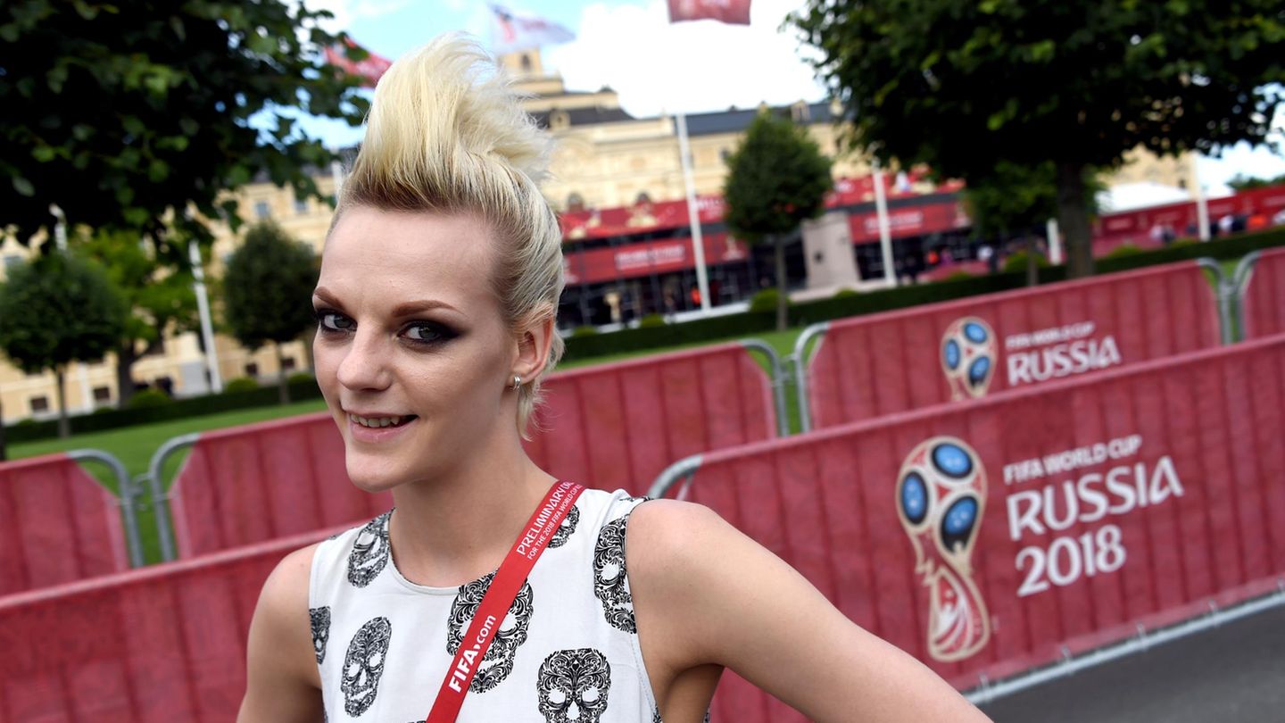 Eine Frau mit blonden, hochtoupierten Haaren lächelt vor einem Plakat mit dem Logo der Fußball-WM 2018 in Russland
