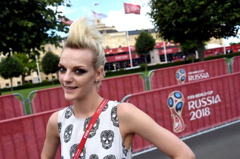 Eine Frau mit blonden, hochtoupierten Haaren lächelt vor einem Plakat mit dem Logo der Fußball-WM 2018 in Russland