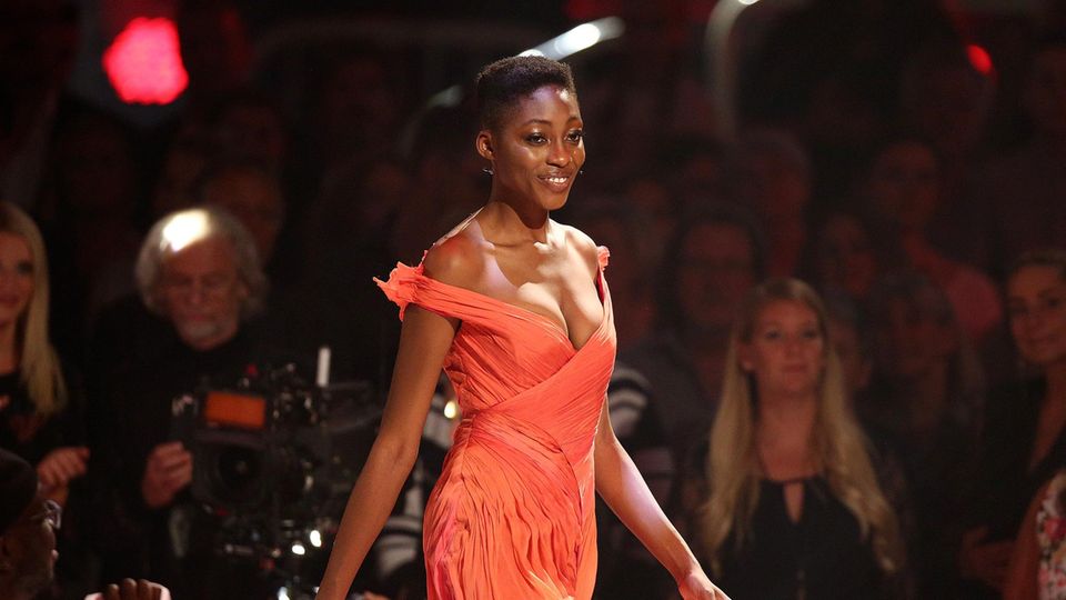 Oluwatoniloba Dreher-Adenuga Ein farbiges Model läuft in einem schulterfreien, apricotfarbenen Kleid über einen Laufsteg im TV-Studio