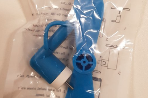 Ein kleiner blauer Ventilator mit USB-Anschluss liegt in einem Plastikbeute auf einer weißen Fläche