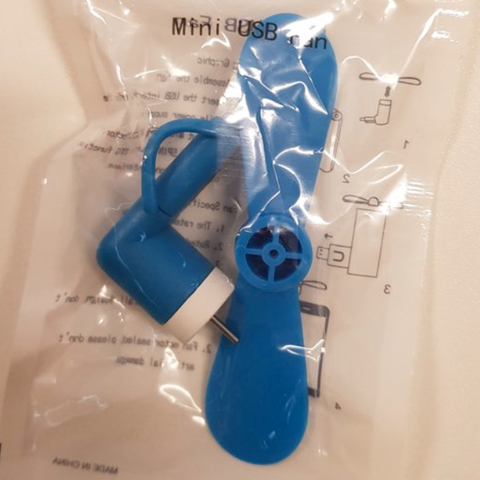 Ein kleiner blauer Ventilator mit USB-Anschluss liegt in einem Plastikbeute auf einer weißen Fläche