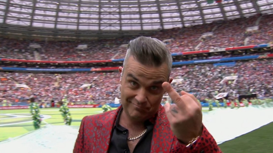Robbie Williams steht im roten Jackett mit Leo-Print auf der Bühne und hält seinen Mittelfinger in die Kamera