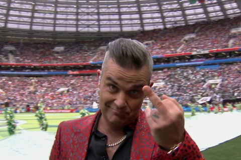 Robbie Williams steht im roten Jackett mit Leo-Print auf der Bühne und hält seinen Mittelfinger in die Kamera