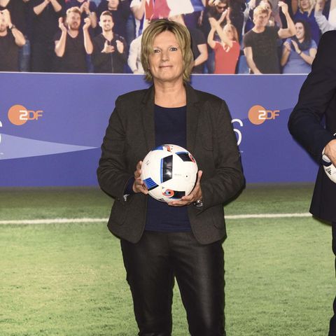 ZDF-Kommentatorin steht im dunklen Blazer und mit Fußball in den Händen in einem TV-Studio