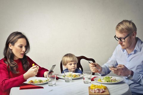 Vater, Mutter, Kind beim Essen, beide Erwachsene halten Smartphones in den Händen