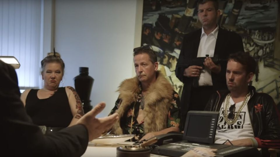 Eine Frau und zwei Männer sitzen an einem Tisch. Hinter ihnen steht ei n Bodyguard mit Gewehr