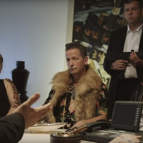 Eine Frau und zwei Männer sitzen an einem Tisch. Hinter ihnen steht ei n Bodyguard mit Gewehr