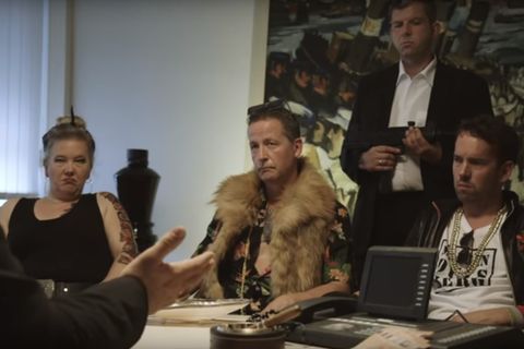 Eine Frau und zwei Männer sitzen an einem Tisch. Hinter ihnen steht ei n Bodyguard mit Gewehr