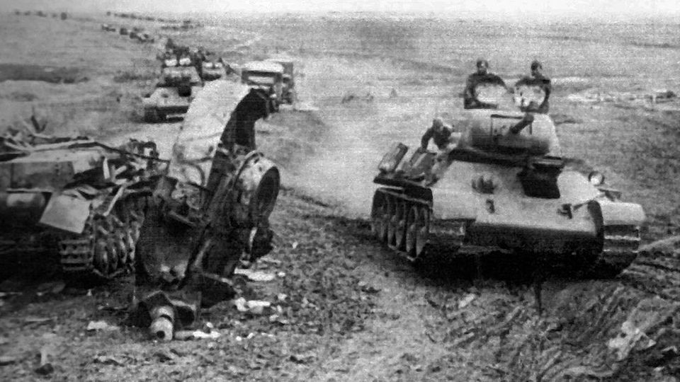 Eine Kolonne T-34 fährt an einem zerstörten Panzer IV vorbei.