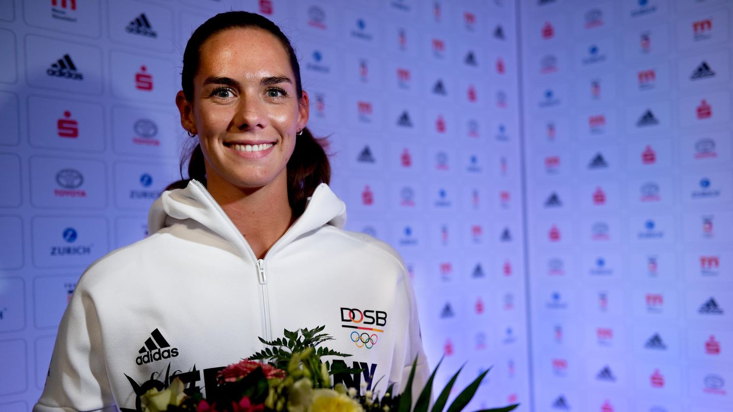 Beachvolleyballerin Kira Walkenhorst steht in weißer Trainingsjacke und mit Blumenstrauß in den Händen vor einer Wand mit Logos