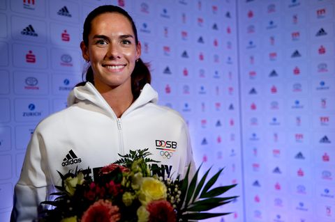 Beachvolleyballerin Kira Walkenhorst steht in weißer Trainingsjacke und mit Blumenstrauß in den Händen vor einer Wand mit Logos