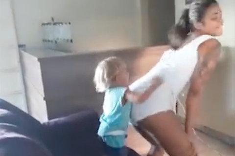 Eine junge Frau in weißem Top und Hotpants twerkt für ihren kleinen Sohn, der auf dem Sofa steht