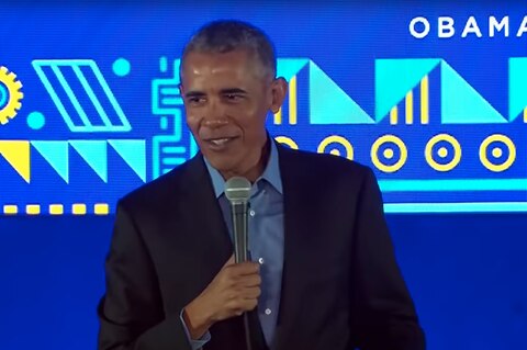 Vor einer blau gemusterten Wand steht Es-US-Präsident Barack Obama auf einer Bühne und hält ein Mikrofon