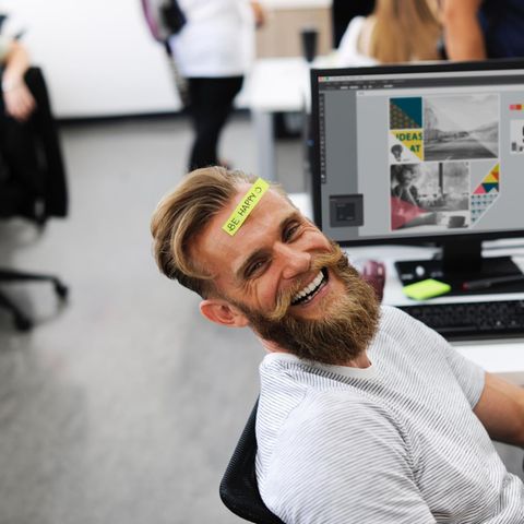 Ein Mann sitzt lachend vor seinem Schreibtisch, auf seiner Stirn ein Post-it mit "Be happy"