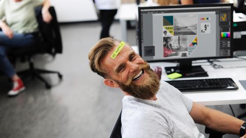 Ein Mann sitzt lachend vor seinem Schreibtisch, auf seiner Stirn ein Post-it mit "Be happy"