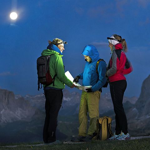 Drei Menschen in Wanderkleidung und mit Kopflampen blicken auf eine Karte. Um sie herum sind Felsen und der Vollmond.