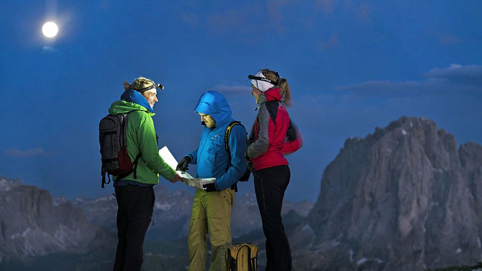 Drei Menschen in Wanderkleidung und mit Kopflampen blicken auf eine Karte. Um sie herum sind Felsen und der Vollmond.