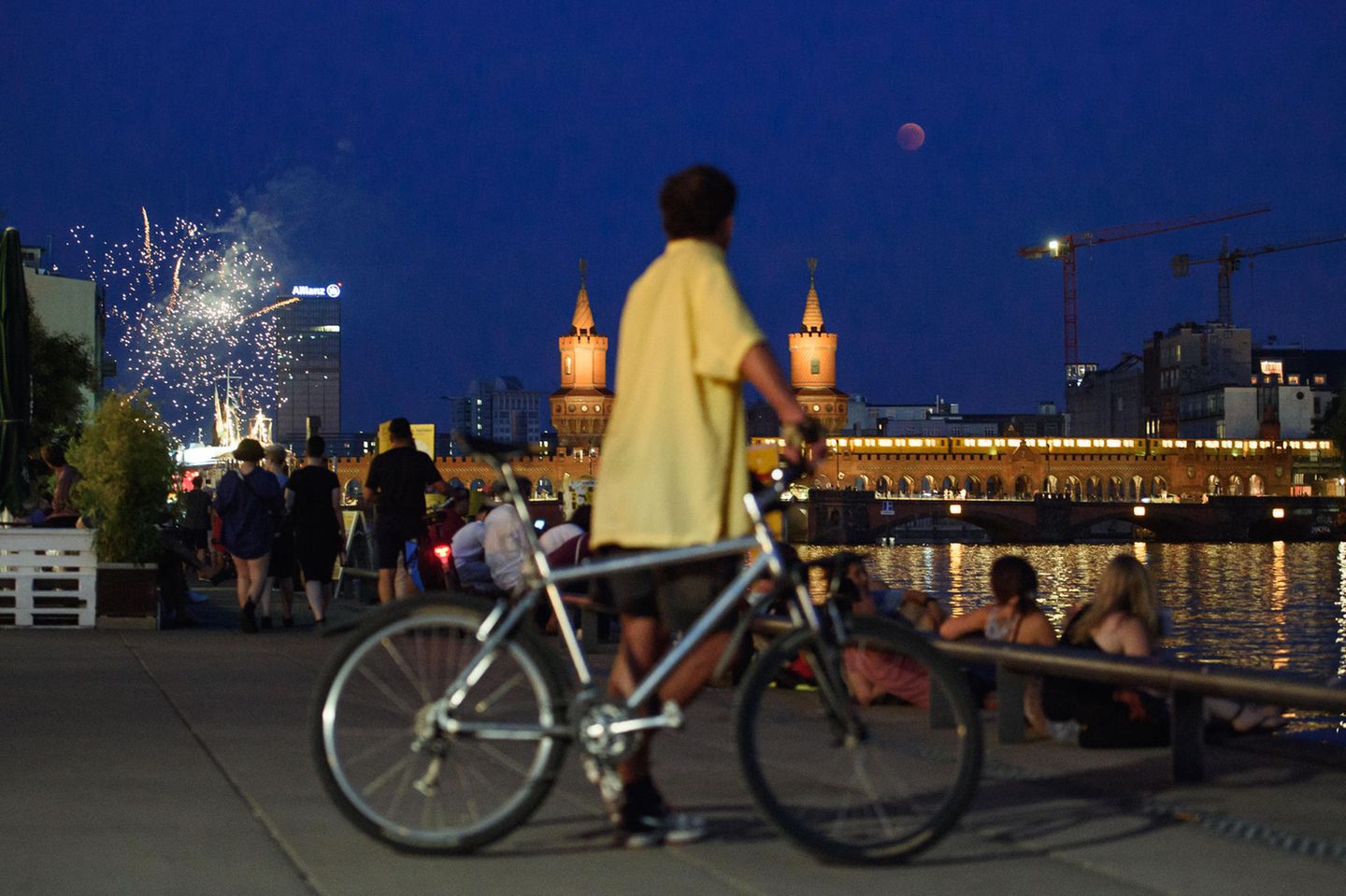 Der Vollmond taucht über der Oberbaumbrücke über der Spree auf, während Berliner sich am Spreeufer ein Feuerwerk anschauen