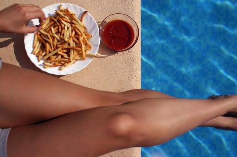 Neben Frauenbeinen am Beckenrand steht ein Teller mit Pommes und daneben eine Schale Ketchup. Eine Hand greift Pommes