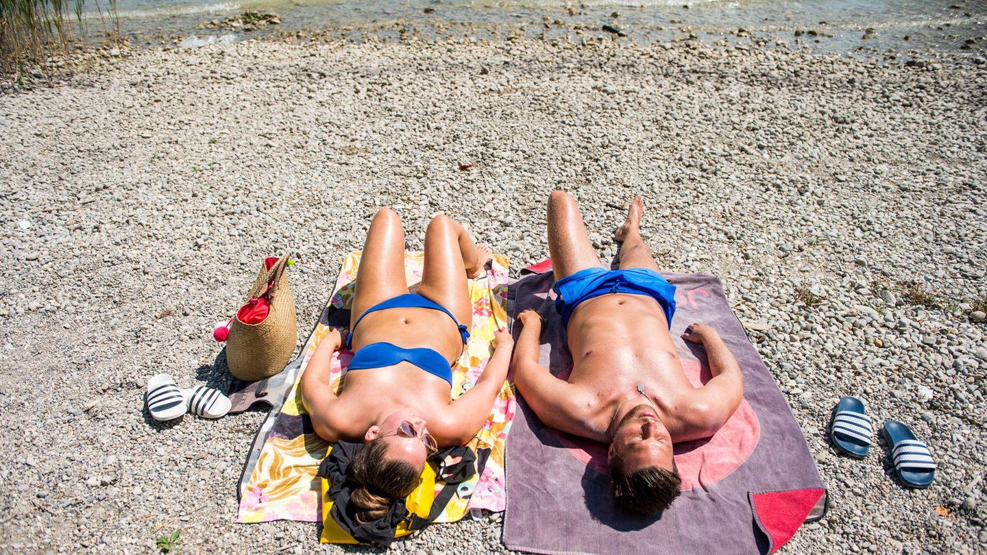 Auf zwei Baedtüchern liegen eine Frau und ein Mann in Badesachen nebeneinander auf einem Kiesstrand. Am oberen Bildrand der See