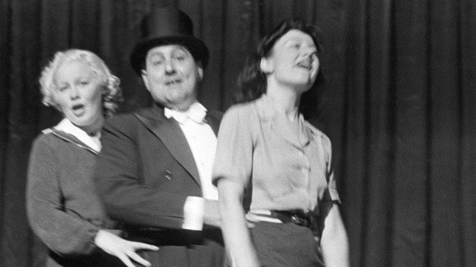Ein Mann mit Zylinder zwischen zwei jungen Frauen auf der Bühne