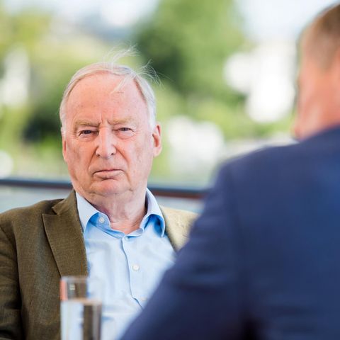 Alexander Gauland sitzt in olivgrünem Jackett und offenem Hemdkragen an einem Tisch. Unscharf im Hintergrund sind Bäume zu sehen