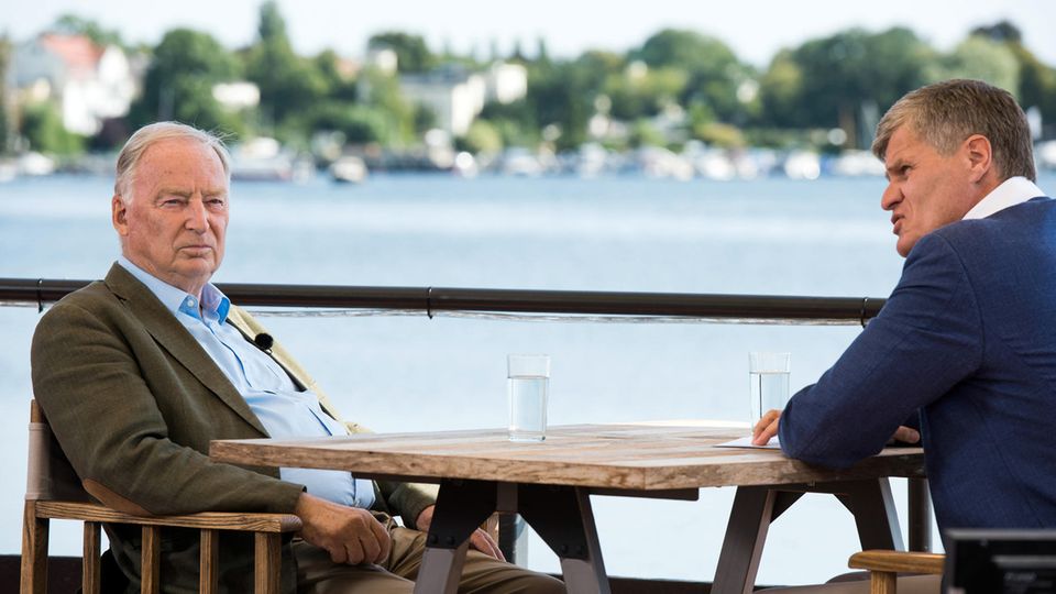 An einem Holztisch sitzen Alexander Gauland von der AfD und Journalist Thomas Walde von ZDF. Im Hintergrund glänzt ein See