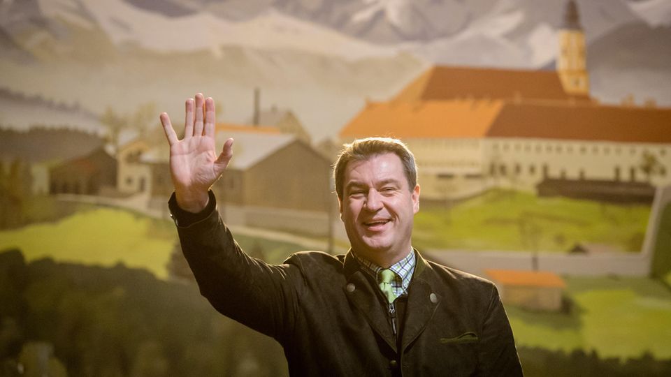 Vor einer gemalten bayrischen Landschaft mit Bergen und Kloster steht Markus Söder im Janker und winkt