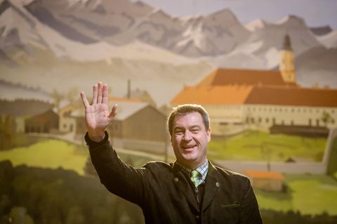 Vor einer gemalten bayrischen Landschaft mit Bergen und Kloster steht Markus Söder im Janker und winkt