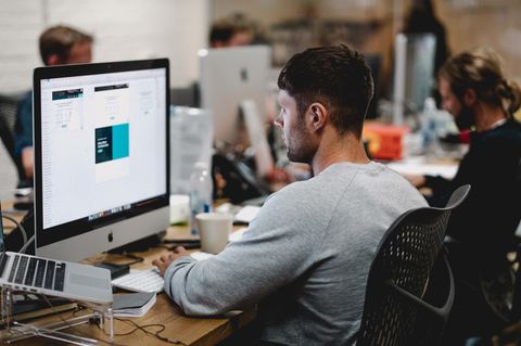 Ein junger Mann sitzt an einem Schreibtisch und arbeitet an einem großen Computer, neben ihm weitere Kollegen