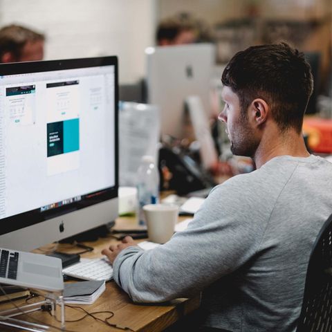 Ein junger Mann sitzt an einem Schreibtisch und arbeitet an einem großen Computer, neben ihm weitere Kollegen