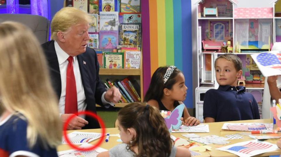 US-Präsident Donald Trump sitzt mit mehreren Kindern an einem Tisch und malt eine US-Flagge aus. Ein Streifen ist blau