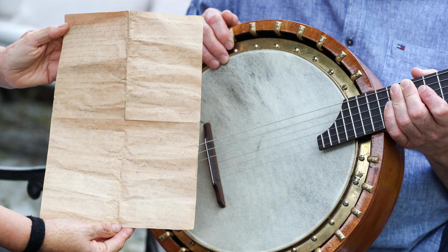 Zwei Hände halten einen vergilbten, oft gefalteten Liebesbrief vor ein altes Banjo, das ein Mann in blauem Hemd hält