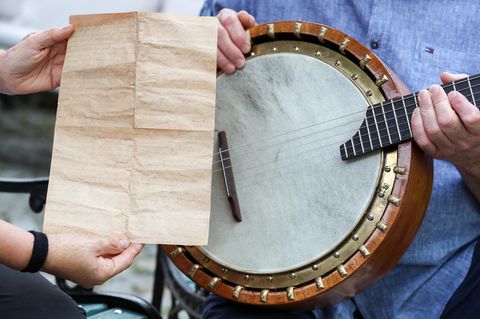 Zwei Hände halten einen vergilbten, oft gefalteten Liebesbrief vor ein altes Banjo, das ein Mann in blauem Hemd hält