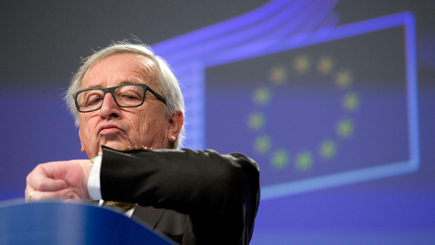 Jean-Claude Juncker schaut im vor einer projezierten EU-Flagge auf die Uhr an seinem linken Handgelenk